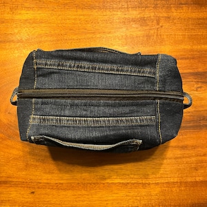 May include: A rectangular dark blue denim pouch featuring a top zipper closure, visible yellow stitching, and integrated denim fabric handles on the sides. The design incorporates horizontal denim panels with reinforced seams and a structured shape.