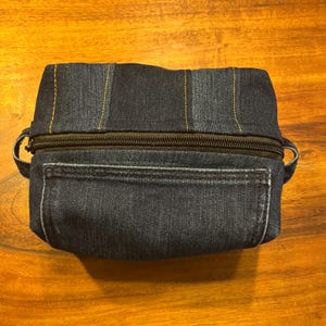 May include: A dark blue denim toiletry bag featuring vertical panels of fabric joined by orange stitching. The bag has a black zippered closure across the top, a rectangular front pocket, and small fabric loops on each side for carrying.