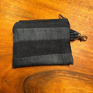 May include: A rectangular zippered pouch crafted from horizontal strips of dark blue and charcoal grey denim fabric. The pouch features a black zipper closure along the top edge and a small fabric tab on the side with a metallic swivel lobster clasp.