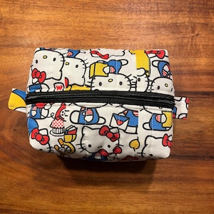 May include: A rectangular quilted fabric pouch featuring a repeating pattern of Hello Kitty characters in white, red, blue, and yellow. The pouch has a black zipper closure running across the top and small fabric tabs on each side of the opening.