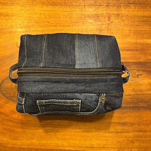 May include: A rectangular pouch crafted from dark blue denim fabric featuring vertical stitched seams, a black zippered closure, and a small side handle. The exterior displays a functional denim pocket with a metallic rivet and visible stitching details.