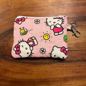 May include: A rectangular pink fabric coin pouch featuring a repeating pattern of Hello Kitty characters, small yellow bees, suns, and white flowers. The pouch has a white zipper closure along the top edge and a silver metal lobster clasp keychain.