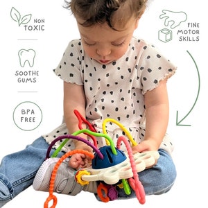 May include: A child is holding a colorful baby toy with curved, flexible tubes in red, yellow, green, and purple. The toy has a white base with a blue center. Text on the image reads: Non Toxic, Soothe Gums, BPA Free, and Fine Motor Skills.