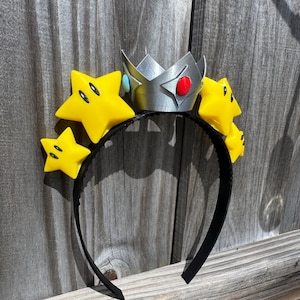 May include: A black headband featuring a silver crown with red and blue accents, and yellow star decorations with black eyes. The crown and stars are attached to the headband, creating a playful accessory.