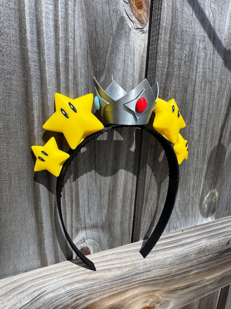 May include: A black headband featuring a silver crown with red and blue accents, and yellow star decorations with black eyes. The crown and stars are attached to the headband, creating a playful accessory.