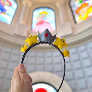 May include: A black headband featuring a silver crown with a red gem and four yellow star accents. The stars have black eyes. The background includes stained glass windows with cartoon characters.