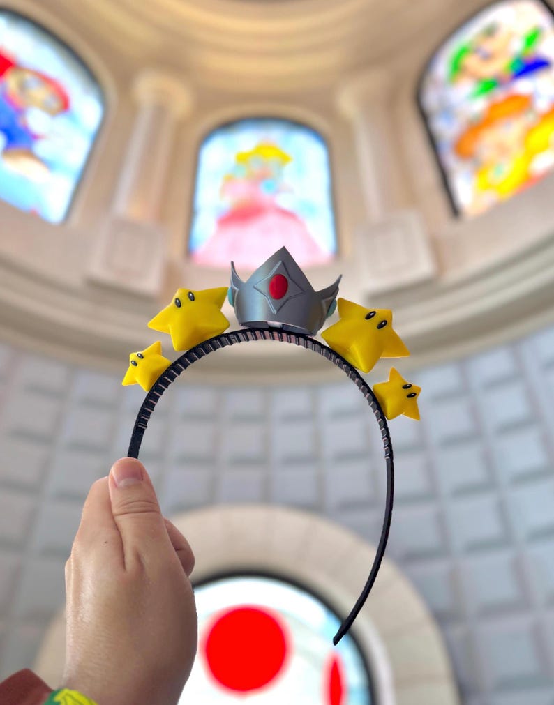May include: A black headband featuring a silver crown with a red gem and four yellow star accents. The stars have black eyes. The background includes stained glass windows with cartoon characters.