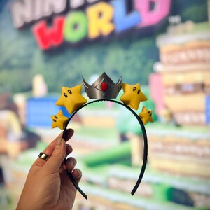 May include: A black headband featuring a silver crown with a red gem and yellow star accents. The stars have black eyes and mouths. The background is a blurred image of a Nintendo World sign.