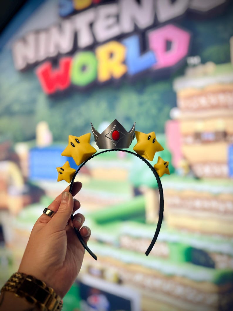 May include: A black headband featuring a silver crown with a red gem and yellow star accents. The stars have black eyes and mouths. The background is a blurred image of a Nintendo World sign.