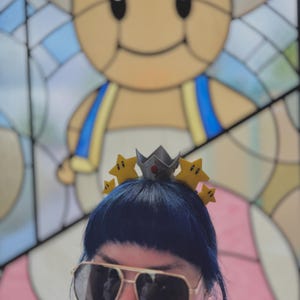 May include: A person wearing a crown-shaped headband with yellow star accents and a silver crown. The person is also wearing gold-rimmed sunglasses with dark lenses. A stained-glass image of a cartoon character is in the background.
