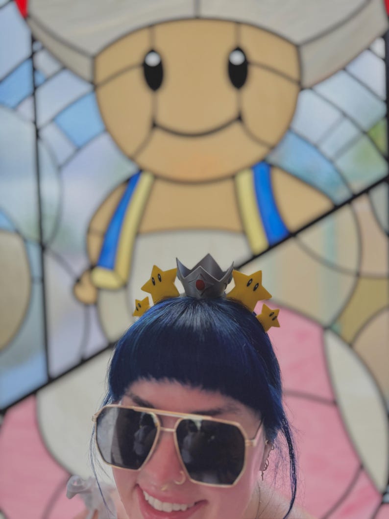 May include: A person wearing a crown-shaped headband with yellow star accents and a silver crown. The person is also wearing gold-rimmed sunglasses with dark lenses. A stained-glass image of a cartoon character is in the background.
