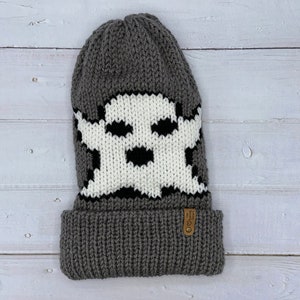 May include: Gray knitted beanie with a white ghost design. The beanie has a fold-over cuff and a brown leather tag with the text "The Knitting  Store" on it.