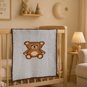 May include: A light blue baby blanket with a brown teddy bear design is draped over a wooden cot. The blanket has a brown border. The nursery has a warm, neutral colour scheme with a teddy bear and a framed print that says "You are so loved."