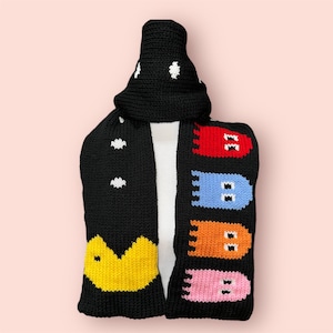 Pode incluir: Cachecol de malha preta com um personagem Pac-Man de um lado e uma fila de fantasmas coloridos do outro. O cachecol é combinado com um chapéu de malha preta com pontos brancos.