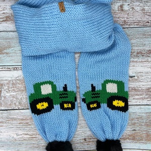 Peut inclure: Une écharpe en tricot bleu clair avec un motif de tracteur vert et jaune et des pompons noirs aux extrémités.