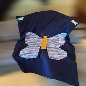 Puede incluir: Una manta azul marino con un gran diseño de mariposa. La mariposa tiene alas coloridas con rayas horizontales en tonos de rosa, azul y verde. El cuerpo de la mariposa es amarillo. Dos pequeños acentos de mariposa están en la manta.