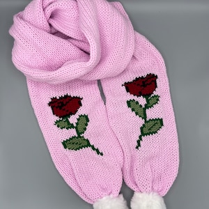 Pode incluir: Uma echarpe de malha rosa com dois desenhos de rosas vermelhas e pompons brancos nas pontas.