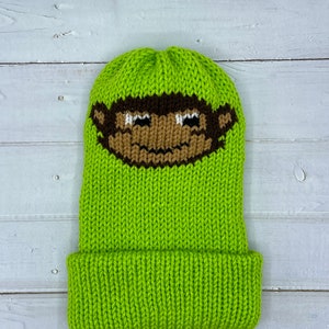 May include: A lime green knitted beanie with a brown monkey face design. The beanie has a fold-over cuff.