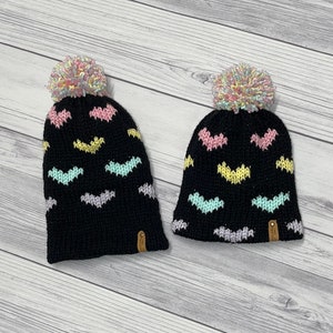May include: Two black knit hats with colorful heart patterns and fluffy pom poms on top.
