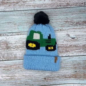 May include: A blue knitted hat with a black pom pom and a green tractor design. The tractor has yellow wheels and black details.