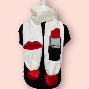 May include: White knitted scarf with red lipstick and lips designs. The scarf has two red pom poms.