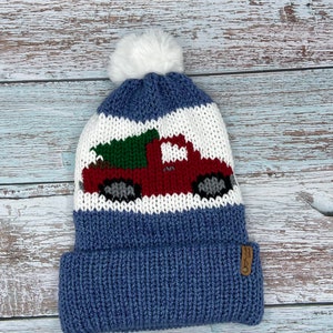 May include: A blue knit beanie with a white pom-pom. The hat features a red truck carrying a green Christmas tree on a white and blue striped background. A small brown tag is sewn on the brim.