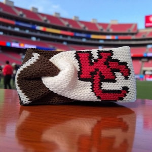 Könnte beinhalten: Ein gestricktes Stirnband mit einem braunen und weißen Muster und einem rot-schwarzen Logo. Das Stirnband besteht aus einem weichen, strukturierten Material und ist für das Tragen um den Kopf konzipiert. Das Logo ist ein stilisiertes Buchstabendesign.