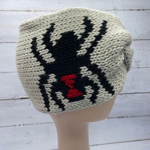 May include: A gray knitted headband with a black spider design. The spider has a red hourglass on its abdomen.