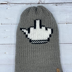 Puede incluir: Gorro de punto gris con un diseño de dedo medio pixelado blanco. El gorro tiene una etiqueta de cuero marrón con el texto "The Knitted Mitten" en ella.