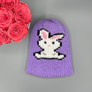 May include: Purple knitted hat with a white bunny design. The bunny has black eyes and pink ears.