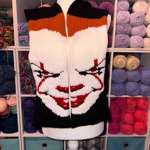 May include: A knitted vest with a zip, depicting a smiling clown face. The vest has a black collar and hem, with orange and white sections. The clown's face is detailed with red lips and black accents.