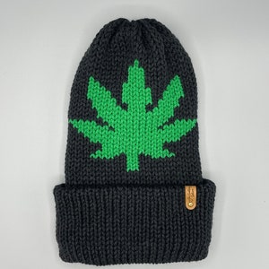 May include: Black knit beanie featuring a large green cannabis leaf design on the front. The beanie has a folded cuff and a small, round, brown tag with text. The hat is made of a thick, textured knit material.