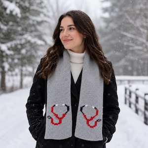 May include: A light gray knit scarf featuring a red and black stethoscope pattern on both ends. The scarf is worn over a white turtleneck sweater and a black coat, with the stethoscope design clearly visible on the front of the gray fabric.