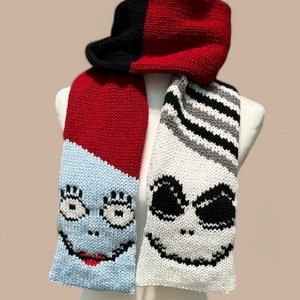 May include: A knitted scarf and hood set featuring character faces. The hood is red and black. One scarf end has a light blue face with red lips and black eyes. The other end has a white face with black eyes and a black smile. The scarf is displayed on a mannequin.
