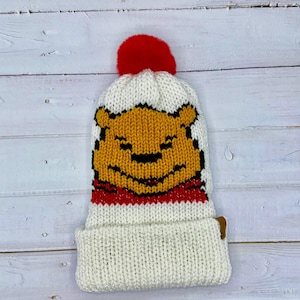 Puede incluir: Gorro de punto blanco con un pompón rojo y un diseño de Winnie de Pooh. El gorro tiene un patrón de rayas rojas y blancas en la parte inferior.