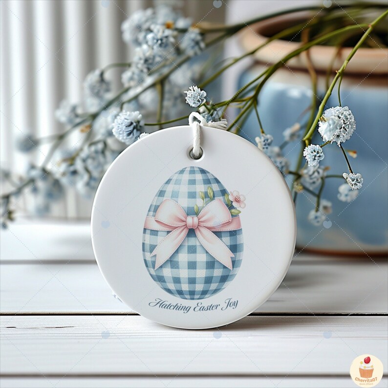 May include: A white ceramic ornament featuring a blue and white gingham egg with a pink bow and the text "Hatching Easter Joy".