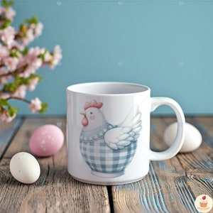 May include: White ceramic mug featuring a blue and white gingham chicken design. The mug has a handle and is resting on a wooden surface with pink and white Easter eggs.