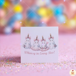 May include: A square card featuring four cartoon ghosts with pink bows and floral accents. The ghosts are attached to clothespins. The text "Whimsy in Every Boo!" is written in pink script below the ghosts. The background is a soft pink with glitter.