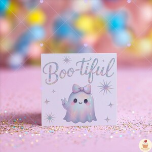 May include: A square card with the text "Boo-tiful" in a holographic font. The card features a cute ghost illustration with a bow, giving a peace sign. The background is a soft pink with glitter.