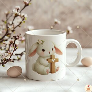 May include: White ceramic mug with a cartoon image of a white bunny holding a wooden cross with a heart on it. The bunny has pink ears and a white flower on its head.