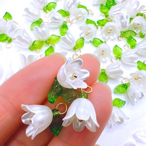 May include: White flower charms with green leaves and gold-colored accents. The charms are in the shape of lily of the valley flowers. The image shows a close-up of the charms, with some held in a hand and others scattered in the background.