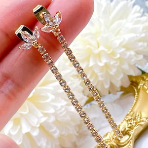 May include: Gold-toned drop earrings with a floral design at the top and a line of small, clear crystals. The earrings are set against a soft, blurred background.