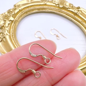 May include: Close-up of gold-colored earring hooks. The hooks are held in a person's hand. The hooks have a curved design with a small loop at the bottom and a coiled wire detail. The background includes a gold-colored decorative frame.