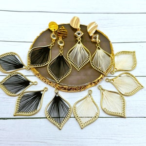 May include: A set of six gold-tone earrings with black and white thread detailing. The earrings are shaped like teardrops and have a delicate, intricate design.