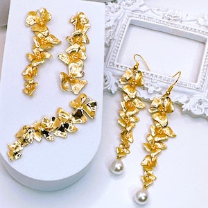 May include: A pair of gold earrings with a floral design. The earrings are long and dangle, with a pearl at the bottom. The earrings are on a white surface.