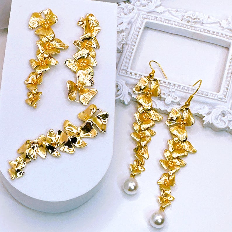 May include: A pair of gold earrings with a floral design. The earrings are long and dangle, with a pearl at the bottom. The earrings are on a white surface.