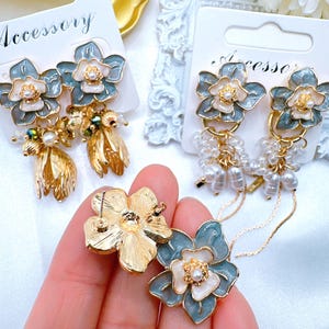 May include: A pair of delicate floral earrings with a light blue and white enamel finish. The earrings feature a flower design with a pearl center and dangle from a gold chain with pearl accents. The earrings are displayed on a white card with the word "Accessory" printed on it.