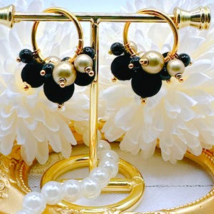 May include: A pair of gold hoop earrings with black and gold beads. The earrings are displayed on a gold stand with a pearl necklace.