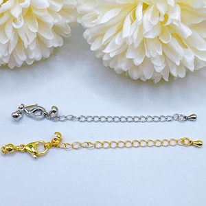 May include: Two necklace extenders, one silver-colored and one gold-colored, are displayed against a soft white background. Each extender features a lobster clasp, a chain of small circular links, and a decorative end. The silver extender has a small ball at the clasp.