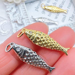 May include: Close-up of gold and silver fish-shaped charms with a textured scale design. Each charm has a small loop for attaching to a necklace or bracelet. The gold charm has a small clear gem on its head.